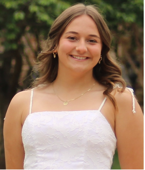Megan Weilmuenster Portrait of Megan Weilmuenster with shoulder-length brown hair, wearing a white sleeveless dress, small earrings, and a gold necklace. She is standing outdoors in front of a blurred green background.